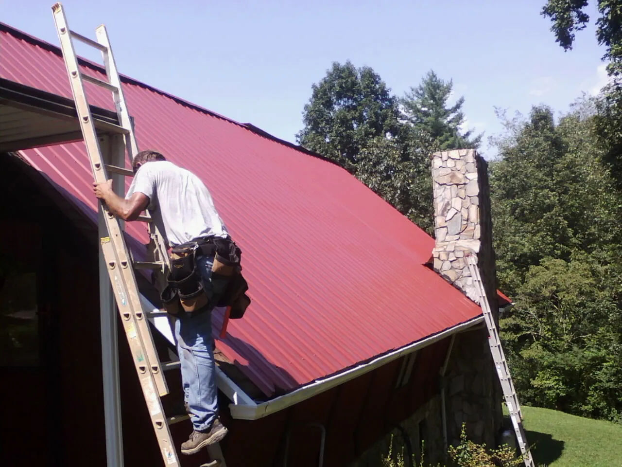 Roofer on a ladder beside a red metal roof and stone chimney — Soffit & Fascia Repair by Roofing Brothers in Cliffwood